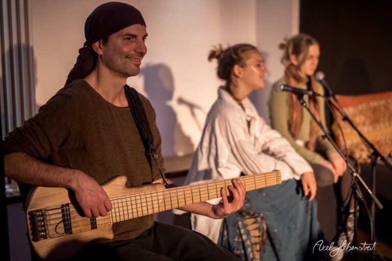 Musiker mit E-Bass spielt bei einer Performance, während Sängerinnen im Hintergrund auf ihre Instrumente warten – © Axel Hebenstreit