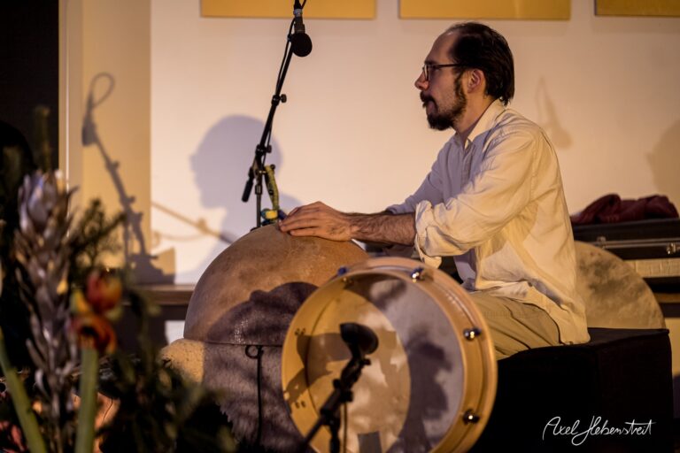 Ein Musiker sitzt auf der Bühne und spielt entspannt auf verschiedenen Trommeln während eines Konzerts – © Axel Hebenstreit