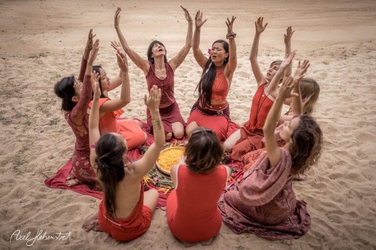 Frauen in roten Kleidern um einen runden Tisch, die mit erhobenen Armen eine kraftvolle Zeremonie im Sand zelebrieren – © Axel Hebenstreit