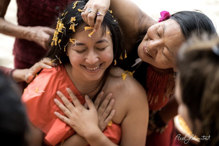 Frauen umarmen eine lächelnde Frau mit Blumenblättern im Haar, in einer festlichen, spirituellen Atmosphäre – © Axel Hebenstreit