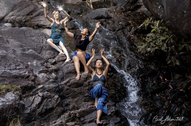 Drei Frauen in Yoga-Pose an einem Wasserfall, umgeben von Felsen und natürlicher Vegetation – © Axel Hebenstreit