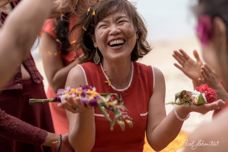 Lachende Frau mit Blumen in den Händen, umgeben von fröhlichen Menschen am Strand – © Axel Hebenstreit