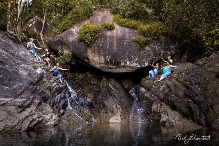 Tanzende Personen an einem felsigen Wasserfall inmitten üppiger Natur – © Axel Hebenstreit