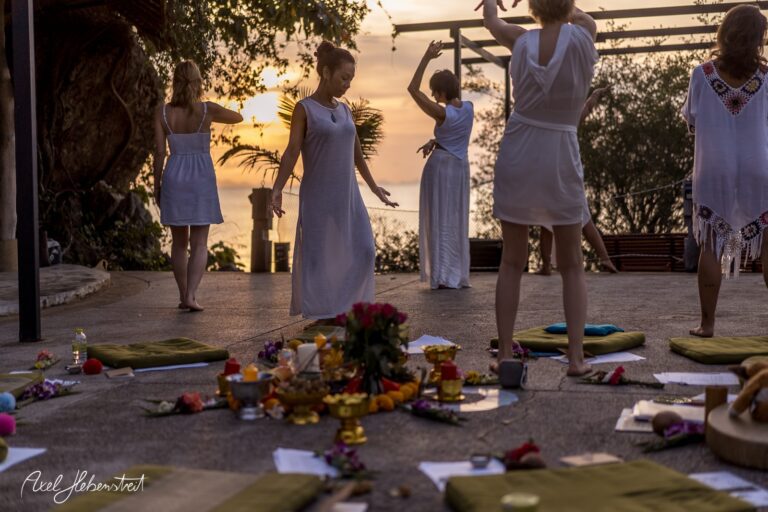 Tanzende Menschen in weißen Kleidern bei Sonnenuntergang umgeben von Blumen und Ritualgegenständen – © Axel Hebenstreit
