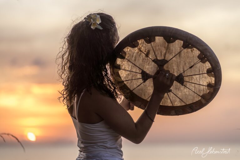 Frau mit zotteligen Haaren und Blüte im Haar spielt eine Trommel vor einem atemberaubenden Sonnenuntergang – © Axel Hebenstreit