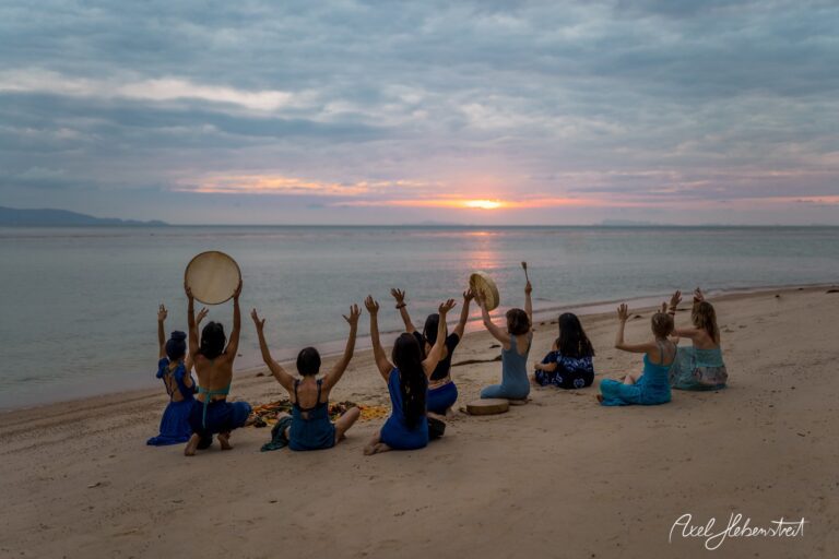 Frauen in blauen Kleidern sitzen am Strand und heben die Hände zum Sonnenuntergang – © Axel Hebenstreit