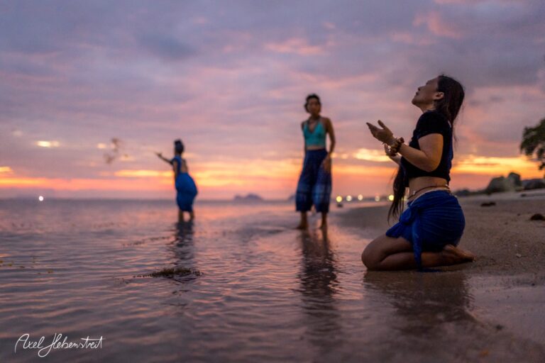 Drei Personen in blauer Kleidung interagieren am Wasser bei Sonnenuntergang, eine kniet betend im Vordergrund. – © Axel Hebenstreit