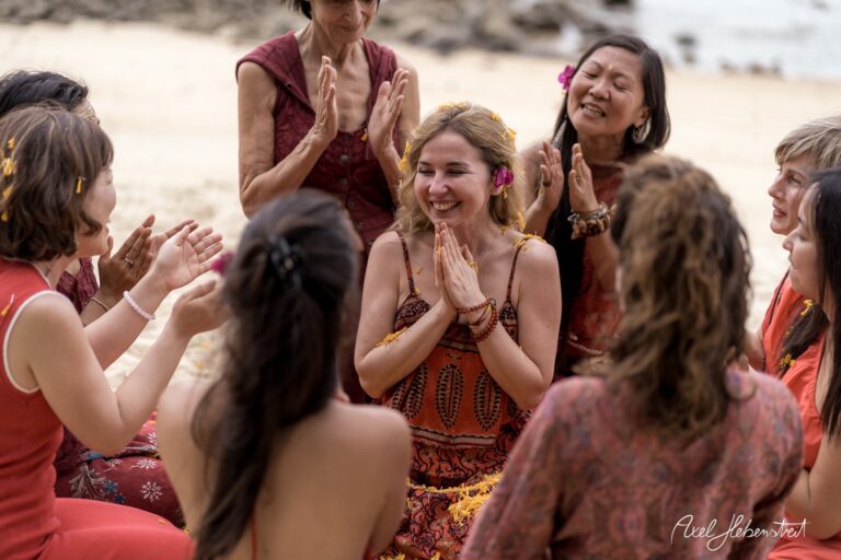 Frauen sitzen im Kreis am Strand und feiern gemeinsam, während eine von ihnen lächelnd im Mittelpunkt steht – © Axel Hebenstreit