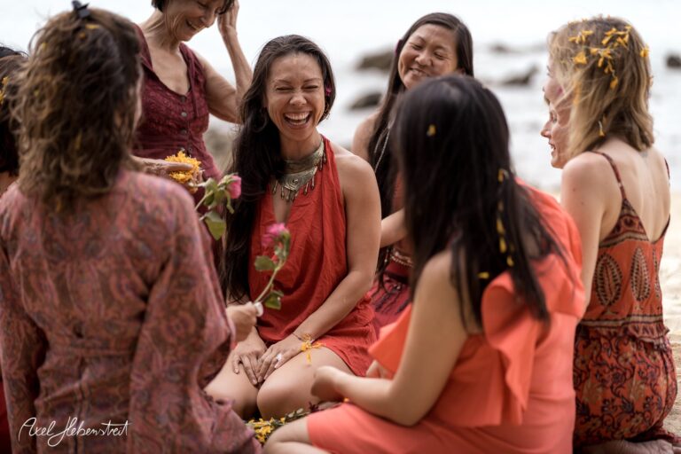 Gruppe fröhlicher Frauen in roten Kleidern sitzt am Strand und teilt einen herzlichen Moment voller Lachen. – © Axel Hebenstreit