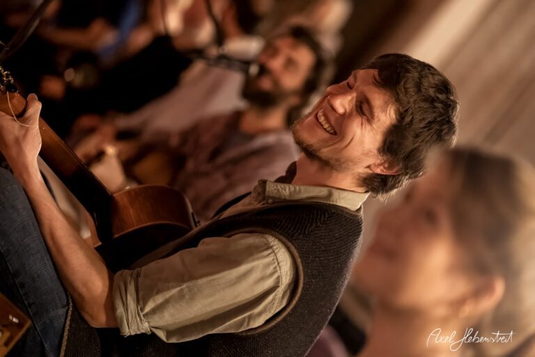 Lächelnder Musiker mit Gitarre, umgeben von einer entspannten Atmosphäre während eines Konzerts – © Axel Hebenstreit