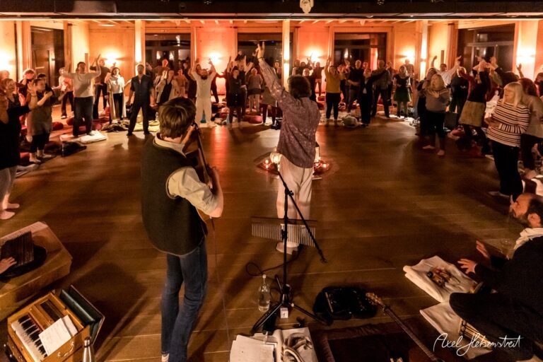 Eine Gruppe von Menschen in einem warm beleuchteten Raum steht zur Musik auf, umgeben von Yogamatten und Instrumenten. – © Axel Hebenstreit
