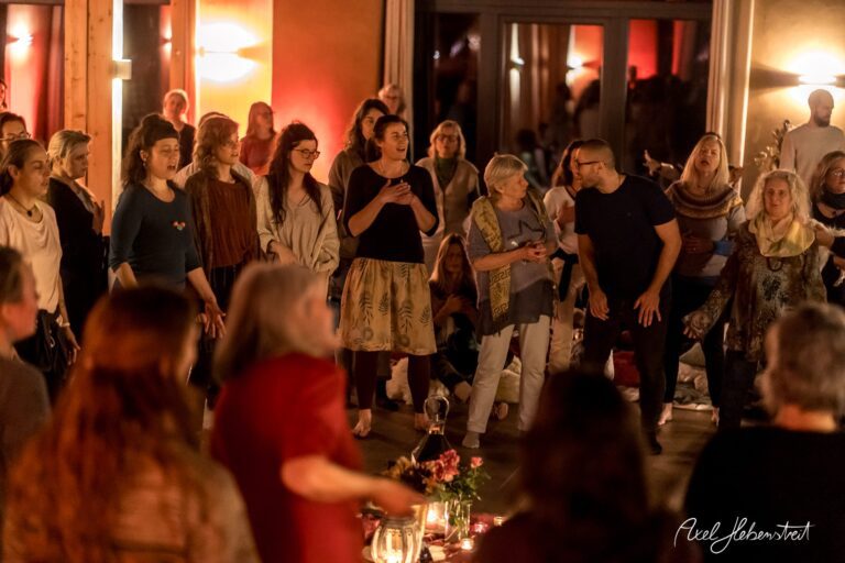 Eine Gruppe von Menschen steht versammelt in einem warmen Raum, während sie an einer gemeinschaftlichen Zeremonie teilnehmen – © Axel Hebenstreit
