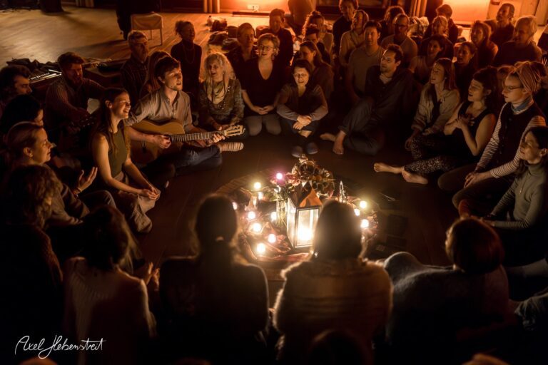 Eine Gruppe von Menschen sitzt im Kreis um Kerzen und Musikinstrumente, während eine intime Atmosphäre entsteht – © Axel Hebenstreit