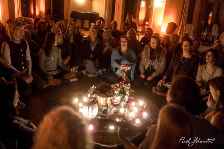 Eine Gruppe von Frauen sitzt kreisförmig um Kerzen in einem stimmungsvollen Raum und praktiziert gemeinsam Yoga oder Meditation. – © Axel Hebenstreit