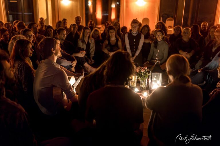 Eine Gruppe von Menschen versammelt sich in einem gemütlichen Raum, begleitet von Musik und Kerzenlicht – © Axel Hebenstreit