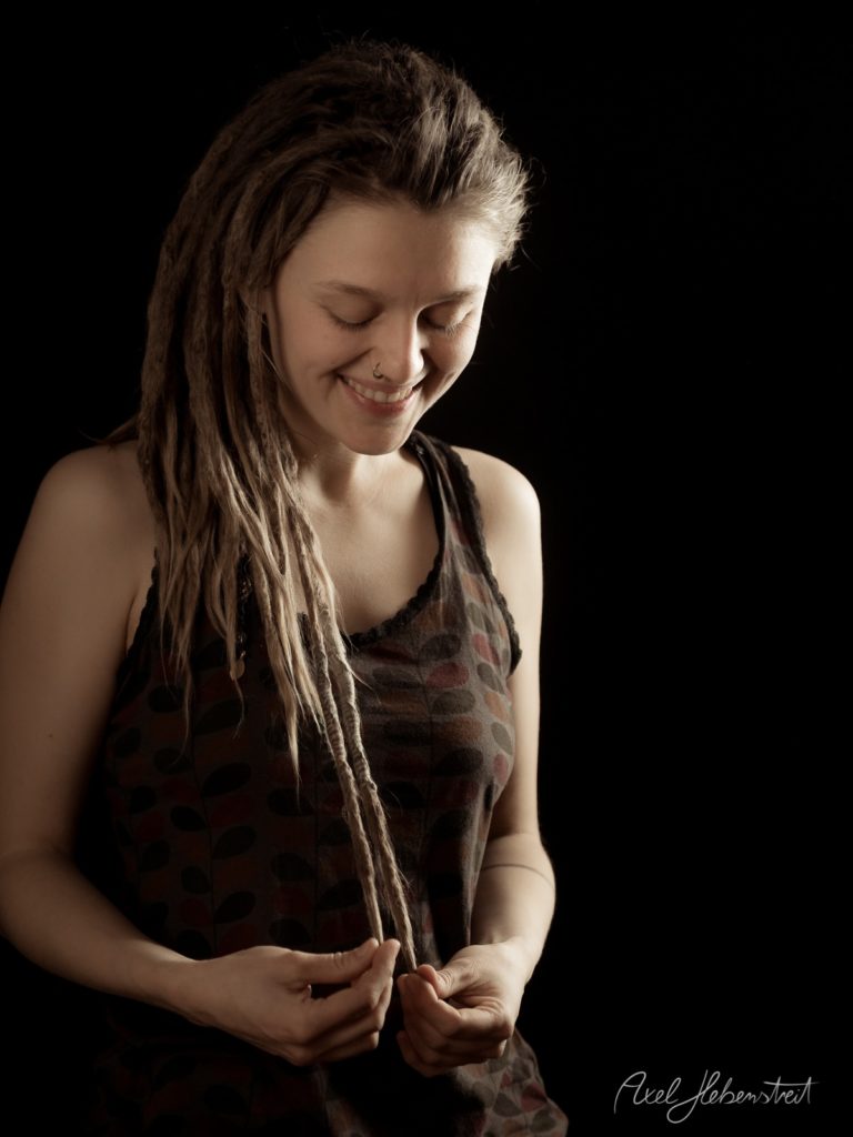 Porträt einer lächelnden Frau mit Dreadlocks in dunklem Tanktop vor schwarzem Hintergrund – © Axel Hebenstreit
