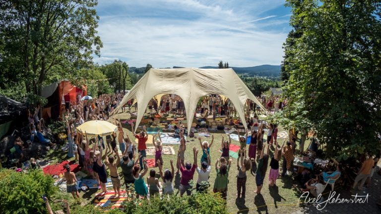 Eine aufgeschlossene Gruppe von Menschen praktiziert Yoga unter einem großen Zelt bei einem Festival in der Natur. – © Axel Hebenstreit