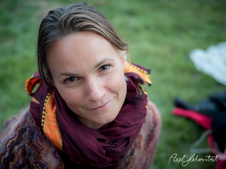 Eine Frau mit einem warmen Lächeln und buntem Schal sitzt im Grünen und strahlt Ruhe aus. – © Axel Hebenstreit