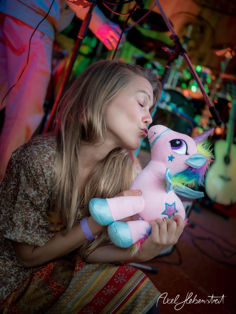 Eine Frau küsst ein plüschiges Einhorn auf einem bunten Festival. – © Axel Hebenstreit