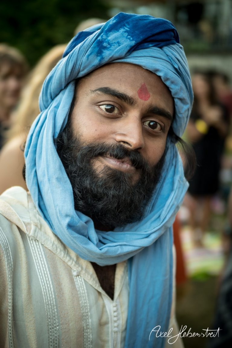 Ein lächelnder Mann mit turbanähnlichem Kopftuch und Beaded-Look blickt freundlich in die Kamera, umgeben von Festivalatmosphäre. – © Axel Hebenstreit