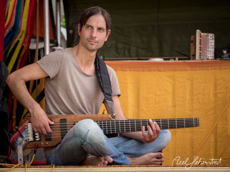 Ein als Musiker verkleideter Mann spielt basssitzt im Freien auf einer Bühne und strahlt Gelassenheit aus. – © Axel Hebenstreit