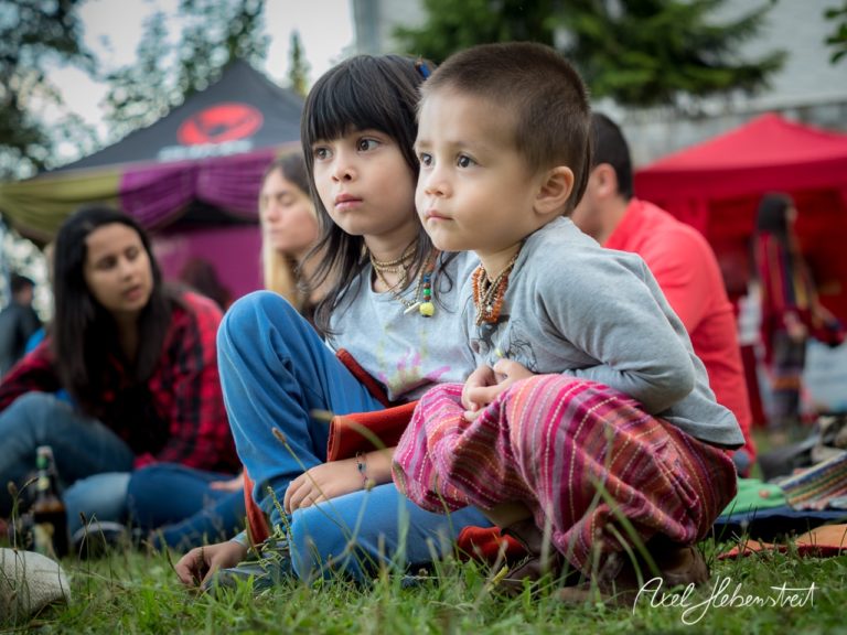 Zwei Kinder sitzen auf dem Boden, freundlich und neugierig, umgeben von einer lebhaften Festivalatmosphäre. – © Axel Hebenstreit