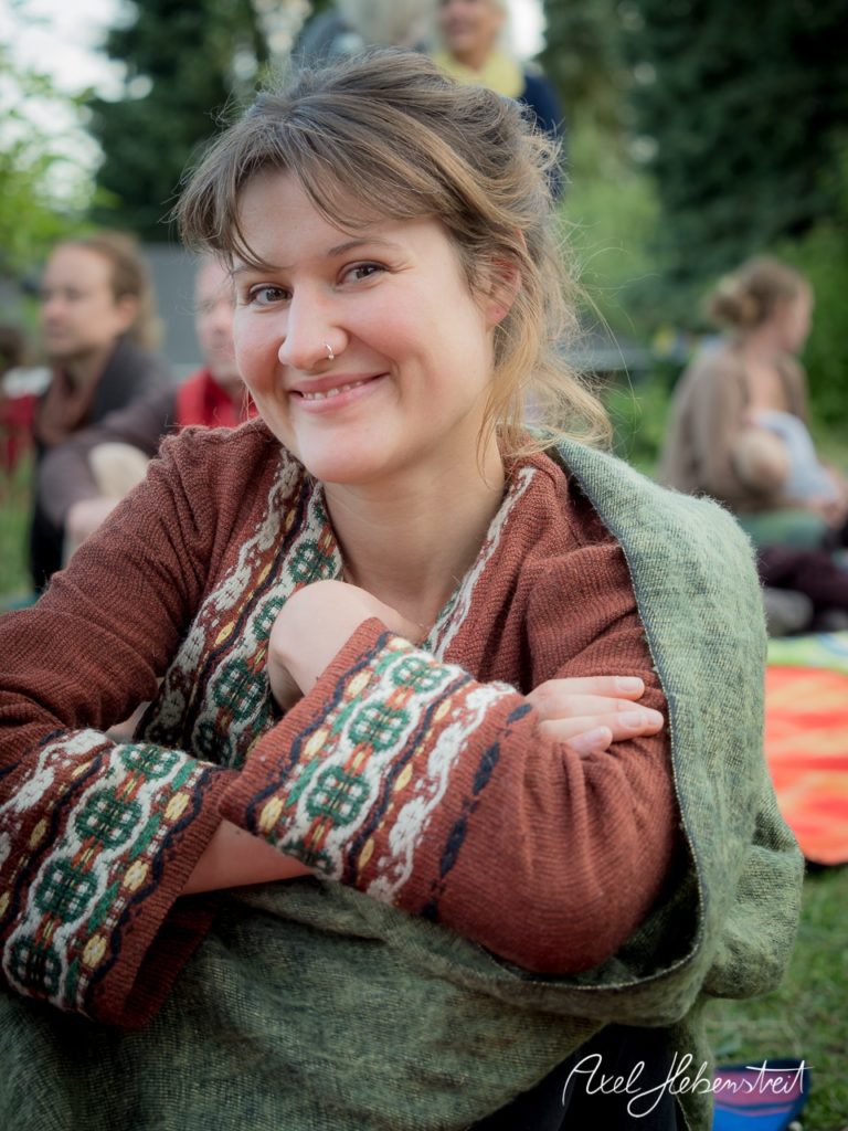 Eine lächelnde Frau in traditioneller Kleidung sitzt entspannt im Freien, umgeben von Natur und anderen Menschen. – © Axel Hebenstreit