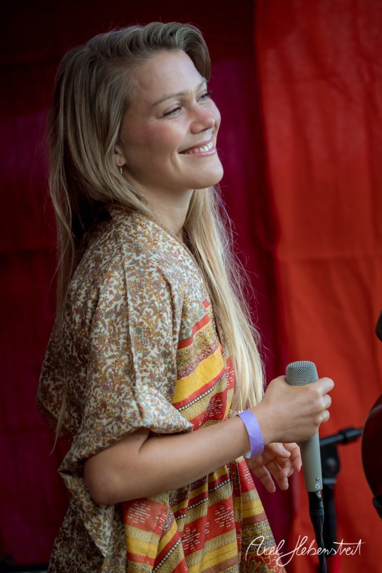 Eine lächelnde Frau mit Mikrofon auf der Bühne, umgeben von farbenfrohen Vorhängen bei einem Festival. – © Axel Hebenstreit