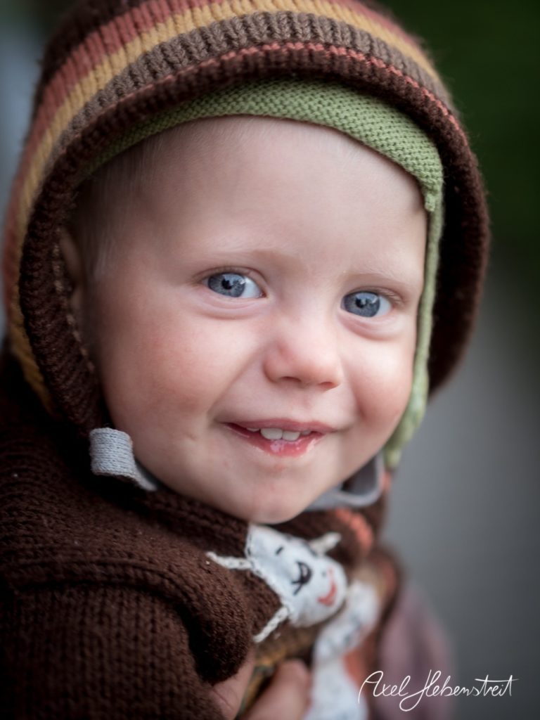 Lächelndes Kleinkind mit blauen Augen in bunter Strickjacke, aufgenommen im Freien. – © Axel Hebenstreit