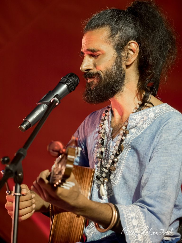Ein Musiker mit Gitarre und lebhaftem Ausdruck spielt auf einer Bühne, umgeben von rotem Licht. – © Axel Hebenstreit