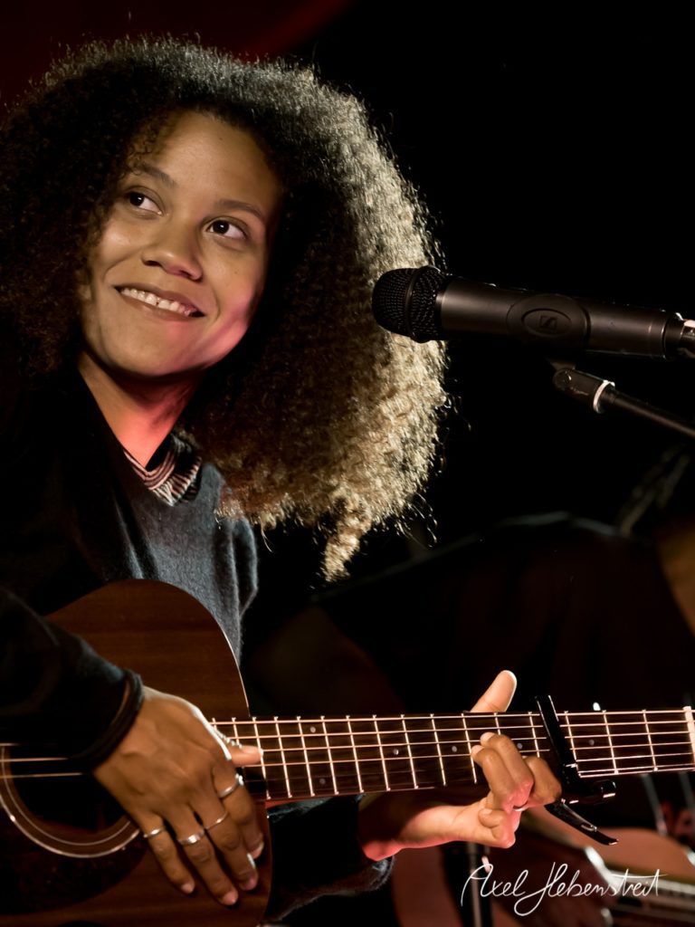 Eine Musikerin mit lockigem Haar spielt mit Freude akustische Gitarre auf einer Bühne. – © Axel Hebenstreit