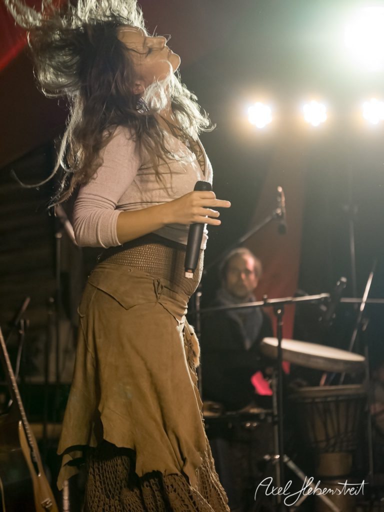 Eine Sängerin mit wallendem Haar performt leidenschaftlich auf der Bühne, während ein Drummer im Hintergrund spielt. – © Axel Hebenstreit