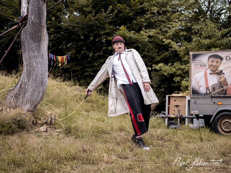 Ein Clown in einem Umhang steht an einem Baum und zieht einen Spielzeugwagen hinter sich her. – © Axel Hebenstreit