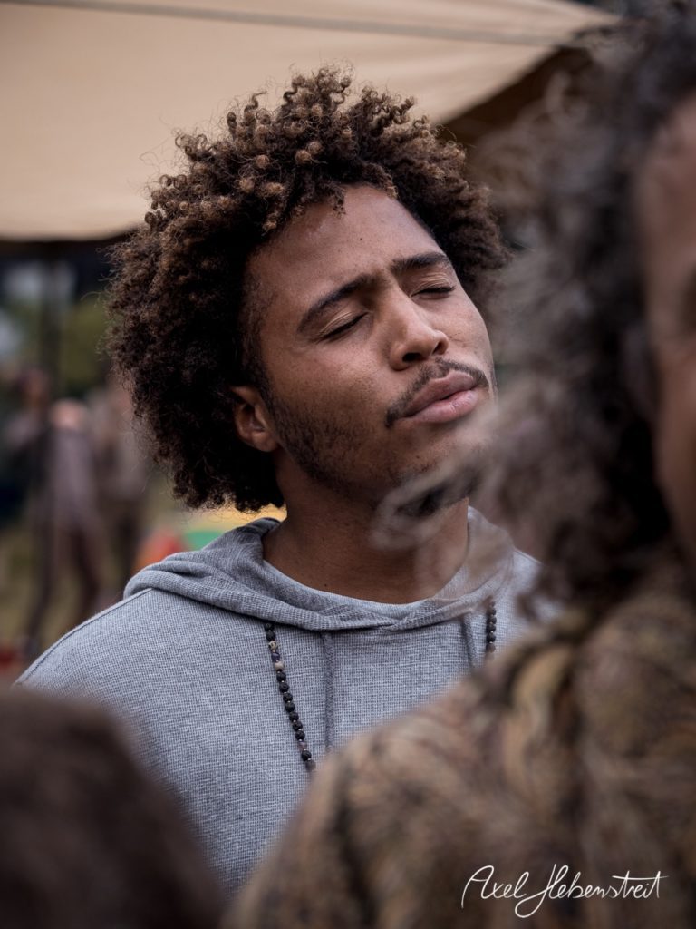 Junger Mann mit lockigem Haar, nachdenklicher Ausdruck, umgeben von Festivalatmosphäre. – © Axel Hebenstreit