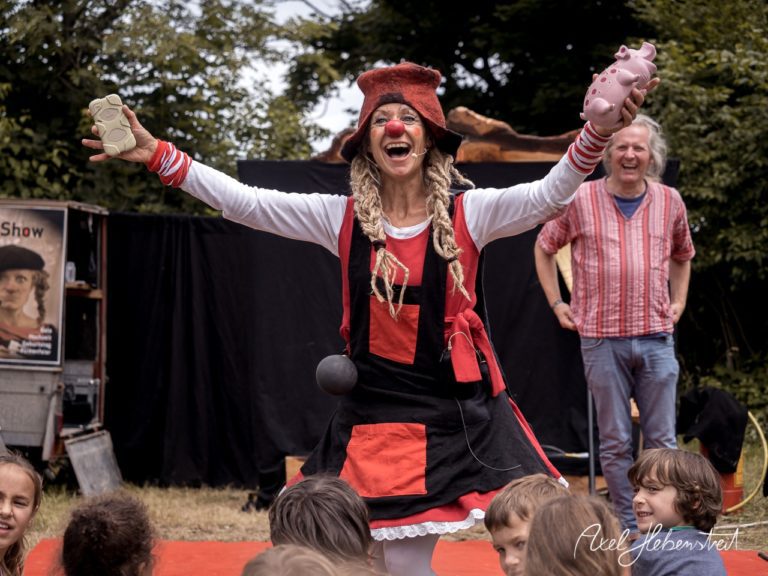 Eine fröhliche Clownin begeistert mit ihrer Performance und Spielzeugfiguren vor einem Publikum im Freien. – © Axel Hebenstreit
