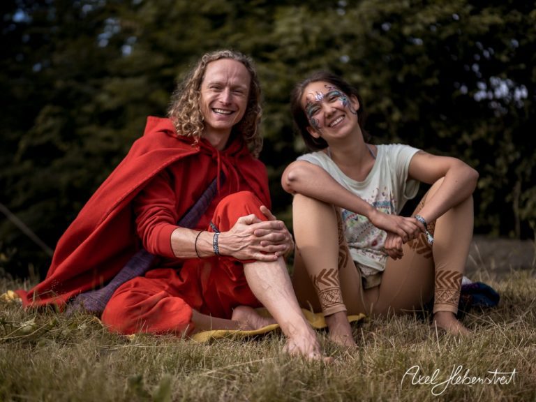 Freudige Momentaufnahme zweier Menschen in bunten Outfits, entspannt sitzend auf einer Wiese bei einem Festival. – © Axel Hebenstreit