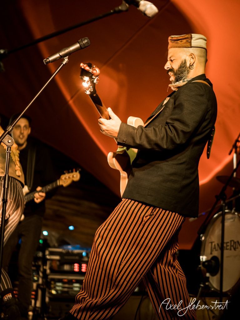 Ein Musiker in traditioneller Kleidung spielt intensiv Gitarre auf einer Bühne während eines Konzerts. – © Axel Hebenstreit