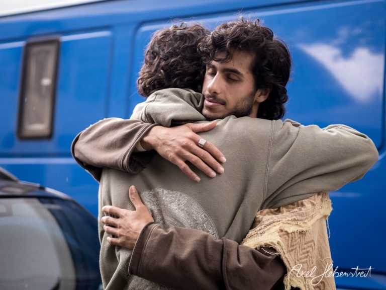 Zwei Männer umarmen sich herzlich vor einem blauen Hintergrund, was Nähe und Verbundenheit ausstrahlt. – © Axel Hebenstreit