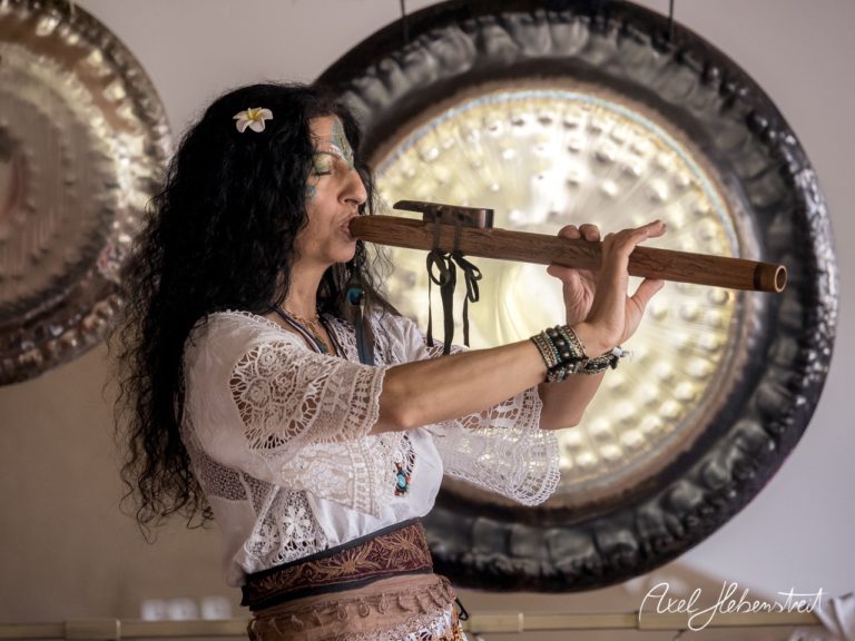 Eine Frau spielt konzentriert eine Flöte in einem harmonischen Raum mit großen Klangschalen im Hintergrund. – © Axel Hebenstreit