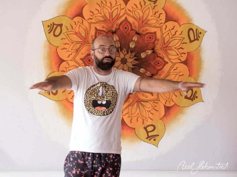 Ein Mann in einem T-Shirt zeigt mit offenen Armen in einem Yoga-Studio vor einer Mandala-Wandmalerei. – © Axel Hebenstreit