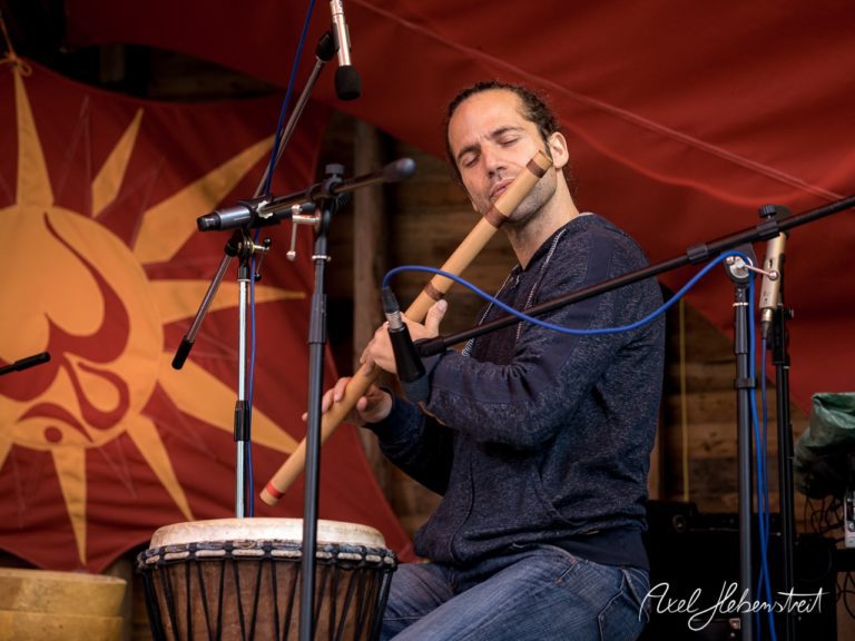 Ein Musiker spielt mit geschlossenen Augen eine Flöte auf der Bühne vor einem bunten Hintergrund. – © Axel Hebenstreit