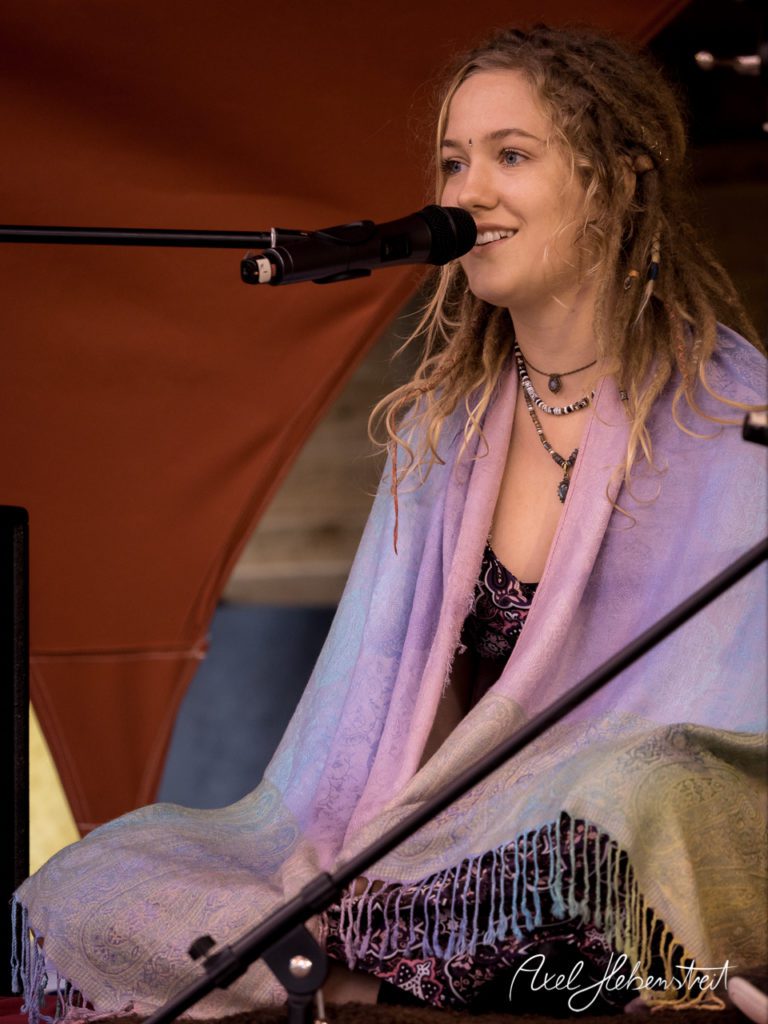 Eine lächelnde junge Frau mit Dreads sitzt auf der Bühne und spricht ins Mikrofon, umgeben von einer warmen Festivalkulisse. – © Axel Hebenstreit