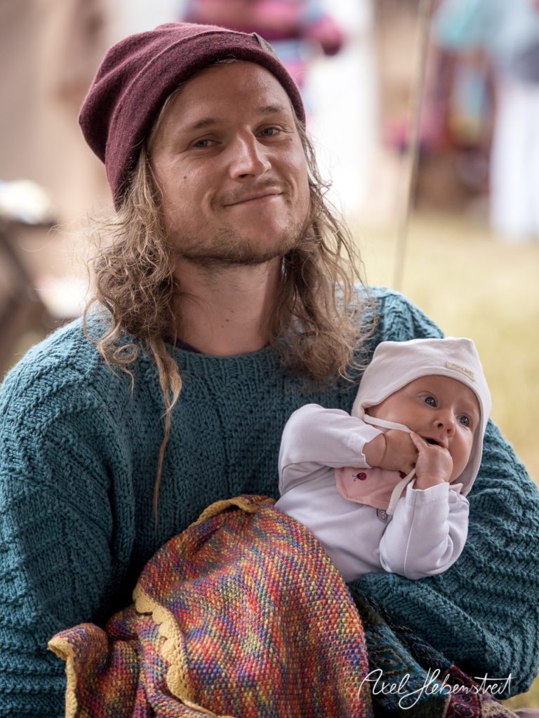 Junger Mann mit langen Haaren und Mütze hält ein Baby auf dem Schoß, umgeben von festivalähnlicher Atmosphäre. – © Axel Hebenstreit