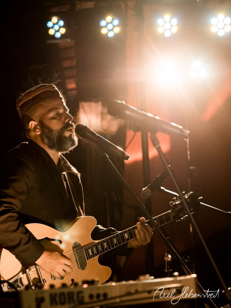 Ein Musiker mit E-Gitarre singt leidenschaftlich auf der Bühne, umgeben von stimmungsvoller Beleuchtung. – © Axel Hebenstreit