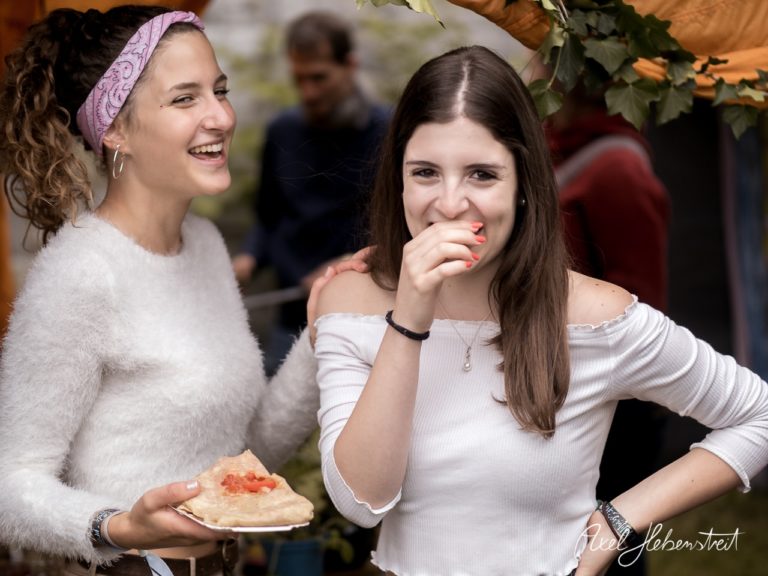 Zwei Frauen lachen fröhlich auf einem Markt, während eine von ihnen ein Stück Essen hält. – © Axel Hebenstreit
