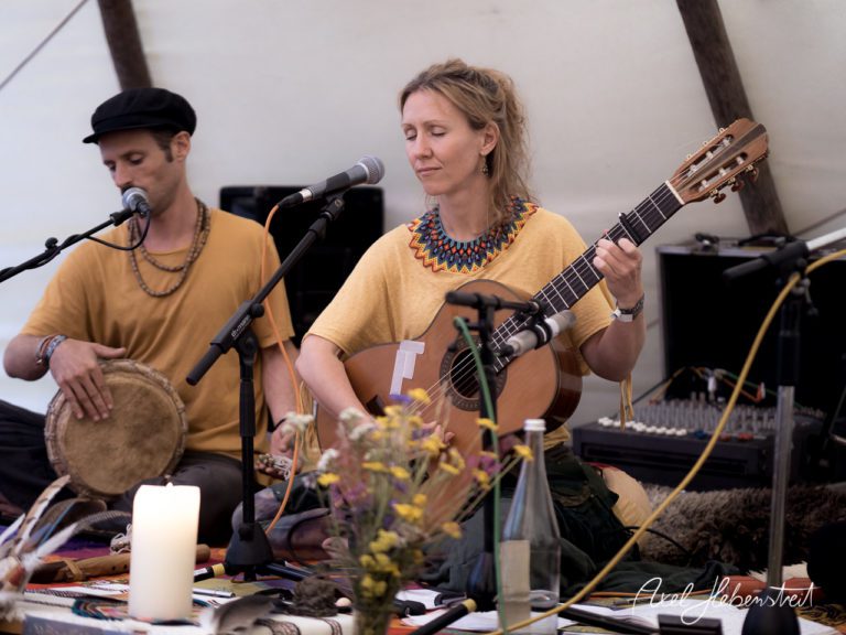 Ein Musiker spielt Percussion, während eine Sängerin mit Gitarre in einer entspannten Festivalatmosphäre auftritt. – © Axel Hebenstreit