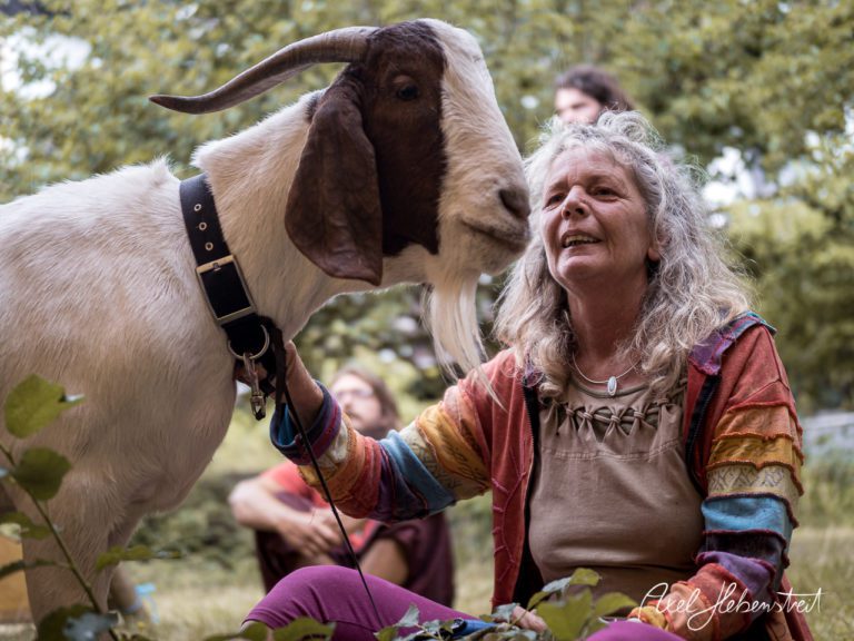 Eine Frau mit lockigem Haar sitzt neben einer Ziege und lächelt, umgeben von grüner Natur. – © Axel Hebenstreit