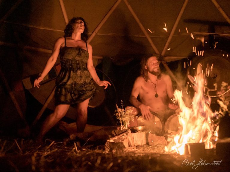 Zwei Menschen tanzen und entspannen sich beim Lagerfeuer in einem Tipi unter warm leuchtenden Holzstrukturen – © Axel Hebenstreit