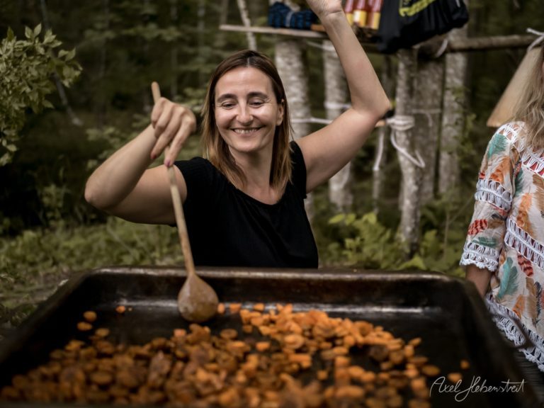 Lachende Frau mit Holzlöffel beim Umrühren von Speisen in schwarzer Schüssel bei Outdoor-Event – © Axel Hebenstreit