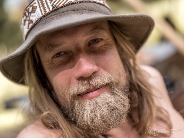Portrait eines Mannes mit langem Haar und Bart, der einen grauen Hut mit gemustertem Band trägt und direkt in die Kamera blickt – © Axel Hebenstreit
